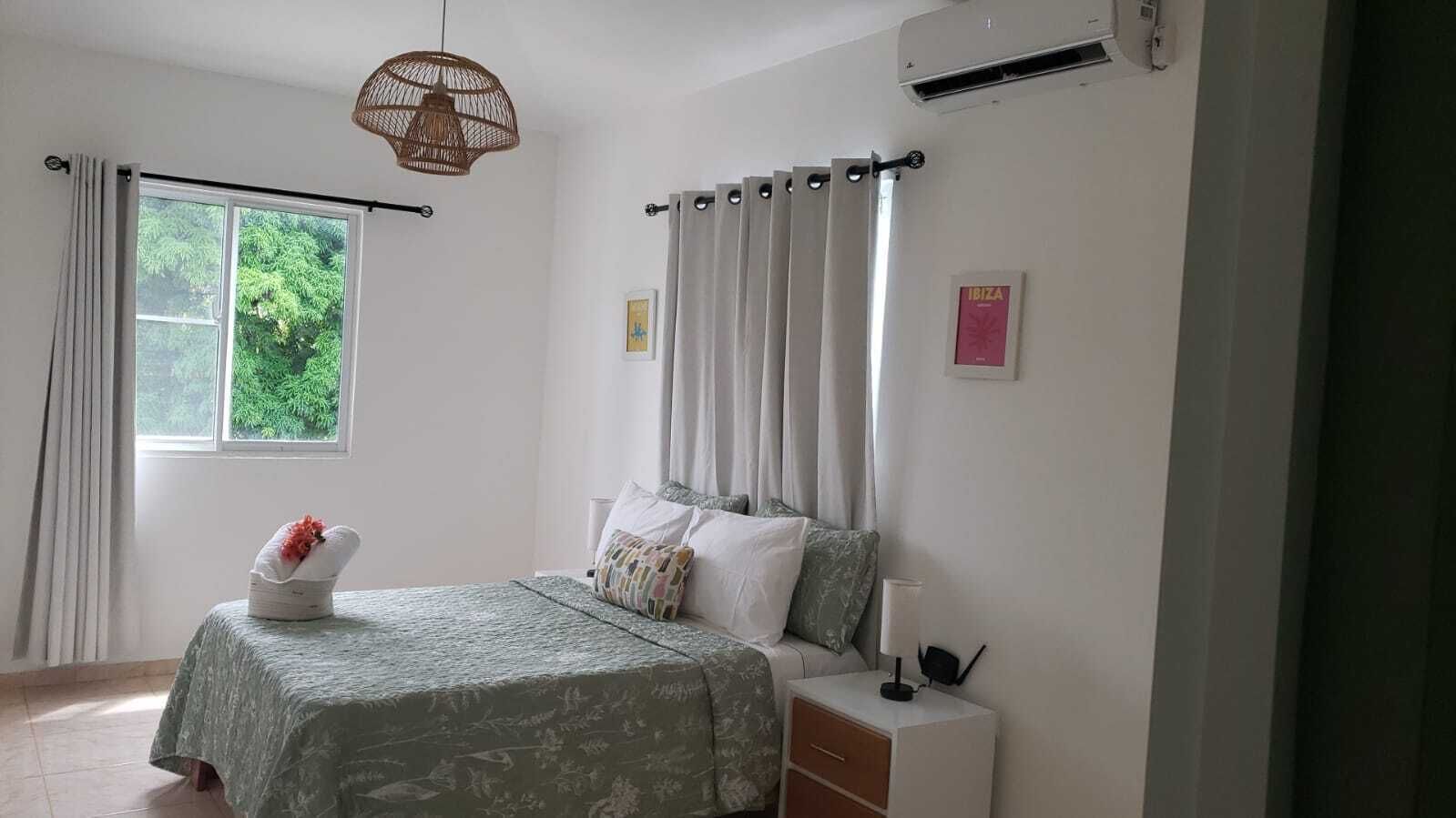 Quarto com cama, mesa de cabeceira, luminária, janela com cortinas e ar condicionado.