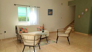 A living room with a sofa, two chairs, a small table, a window with curtains, a staircase, and a few wall-mounted pictures. - Yellow House Río San Juan, Oasis Tropical Pool (Rio San Juan)
