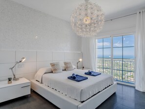 A modern bedroom with a large bed, two bedside tables, a chandelier, and a large window with a city view. - Medussa by Interhome (Dénia)