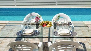 Une table à manger avec des chaises, un panier de fruits et une piscine en arrière-plan.