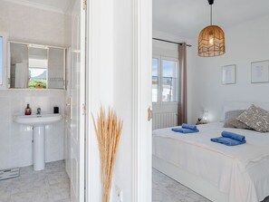 Ein Schlafzimmer mit Bett, Nachttisch, Fenster und einem Badezimmer mit Waschbecken und Spiegel.