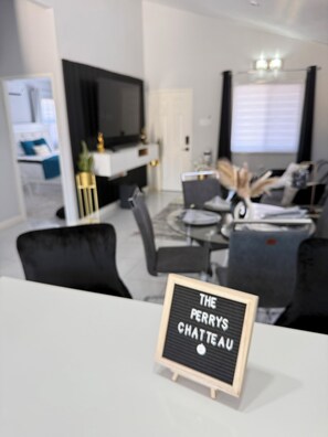 A modern living room with a flat-screen TV, a dining area, and a sign reading 'THE PERRY'S CHATEAU'. - Charming 2-bedroom house with AC in Old Harbour Whim Estates (Old Harbour)