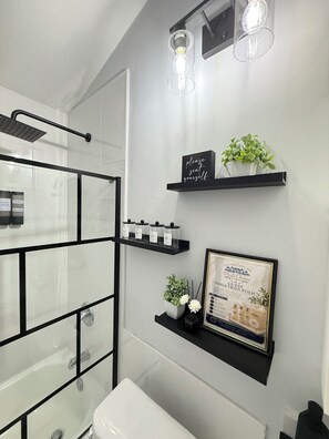 A modern bathroom with a glass shower enclosure, black shelving with toiletries, and a framed guest policy sign. - Charming 2-bedroom house with AC in Old Harbour Whim Estates (Old Harbour)