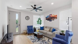 Un salon comprenant un canapé, des fauteuils, une table basse, un téléviseur, un ventilateur de plafond, une horloge et une fenêtre avec des stores.
