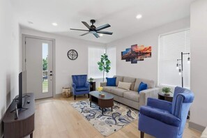 Ein Wohnzimmer mit Sofa, Sesseln, Couchtisch, Fernseher, Deckenventilator, Uhr und einem Fenster mit Jalousien.