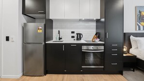 Cozinha compacta com geladeira, fogão e pia, ao lado de uma cama com mesa de cabeceira.