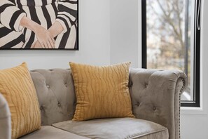 A tufted sofa with two cushions, a framed abstract painting, and a window with a view. - CBD Oasis Studio Pool Sauna & Gym Near Skytower (Auckland CBD)