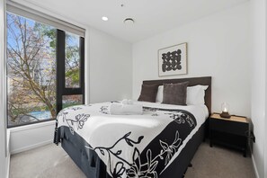 A bedroom with a large window, a bed with a floral bedspread, a nightstand with a lamp, and a framed artwork on the wall. - CBD Oasis Studio Pool Sauna & Gym Near Skytower (Auckland CBD)