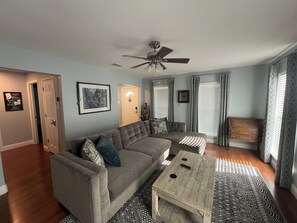 Ein Wohnzimmer mit grauem Sofa, Couchtisch aus Holz, Deckenventilator und einem gemusterten Teppich.