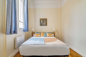 Une chambre à coucher avec un lit, une fenêtre avec des rideaux, un tableau accroché au mur et des lampes de chevet.