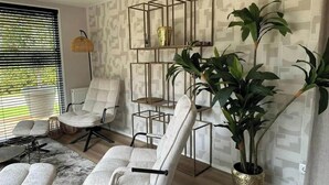 Ein modernes Wohnzimmer mit zwei weißen Sesseln, einer Stehlampe, einer großen Kübelpflanze und einem geometrischen Regalsystem an einer Wand mit Muster.