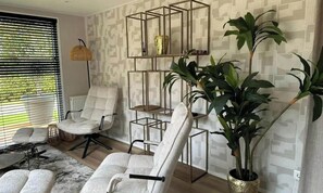 A modern living room with two white armchairs, a floor lamp, a large potted plant, and a geometric shelving unit against a patterned wall. - Chalet de Blauwe Regen With Sun Terrace (Balkbrug)