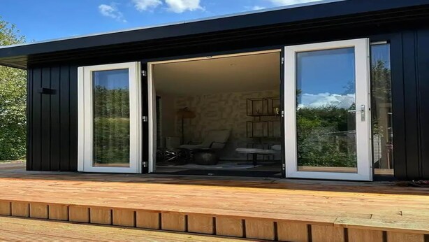 Ein modernes Gebäude mit einer Terrasse, großen Schiebetüren und einem einsehbaren Innenraum mit Möbeln.