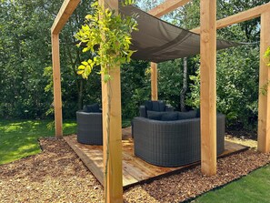 A wooden pergola structure with two wicker sofas under a shaded area, surrounded by a garden with mulch and greenery. - Chalet de Blauwe Regen With Sun Terrace (Balkbrug)