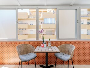 Ein Zimmer mit großem Fenster, zwei Korbstühlen, einem kleinen Tisch mit Flasche und Glas sowie Wandpapier mit Muster.