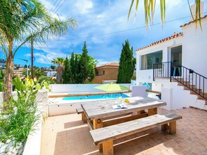 A poolside area with a wooden bench, a table with a book, and a view of a white building with a tiled roof. - Bohemia by Interhome (Calpe/Calp)