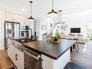 A modern kitchen with stainless steel appliances, a central island, pendant lighting, and a view into the living area. - Desert Oasis • Heated Pool & Spa • Sleeps 8 (Indio)