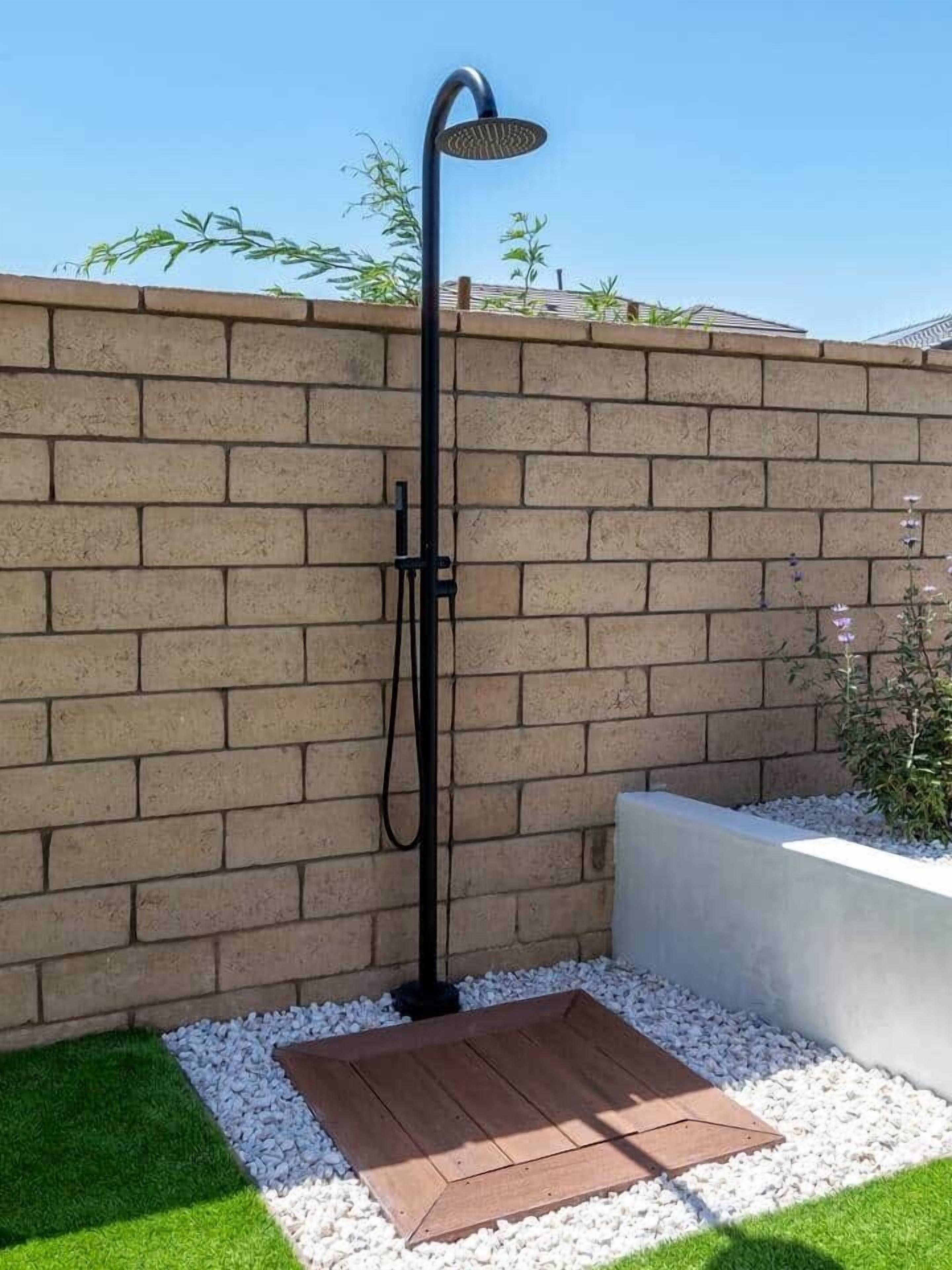 Une douche extérieure moderne, dotée d’un tuyau et d’un pommeau de douche noirs, installée devant un mur de briques et agrémentée d’un petit jardin avec des galets blancs et de l’herbe verte.