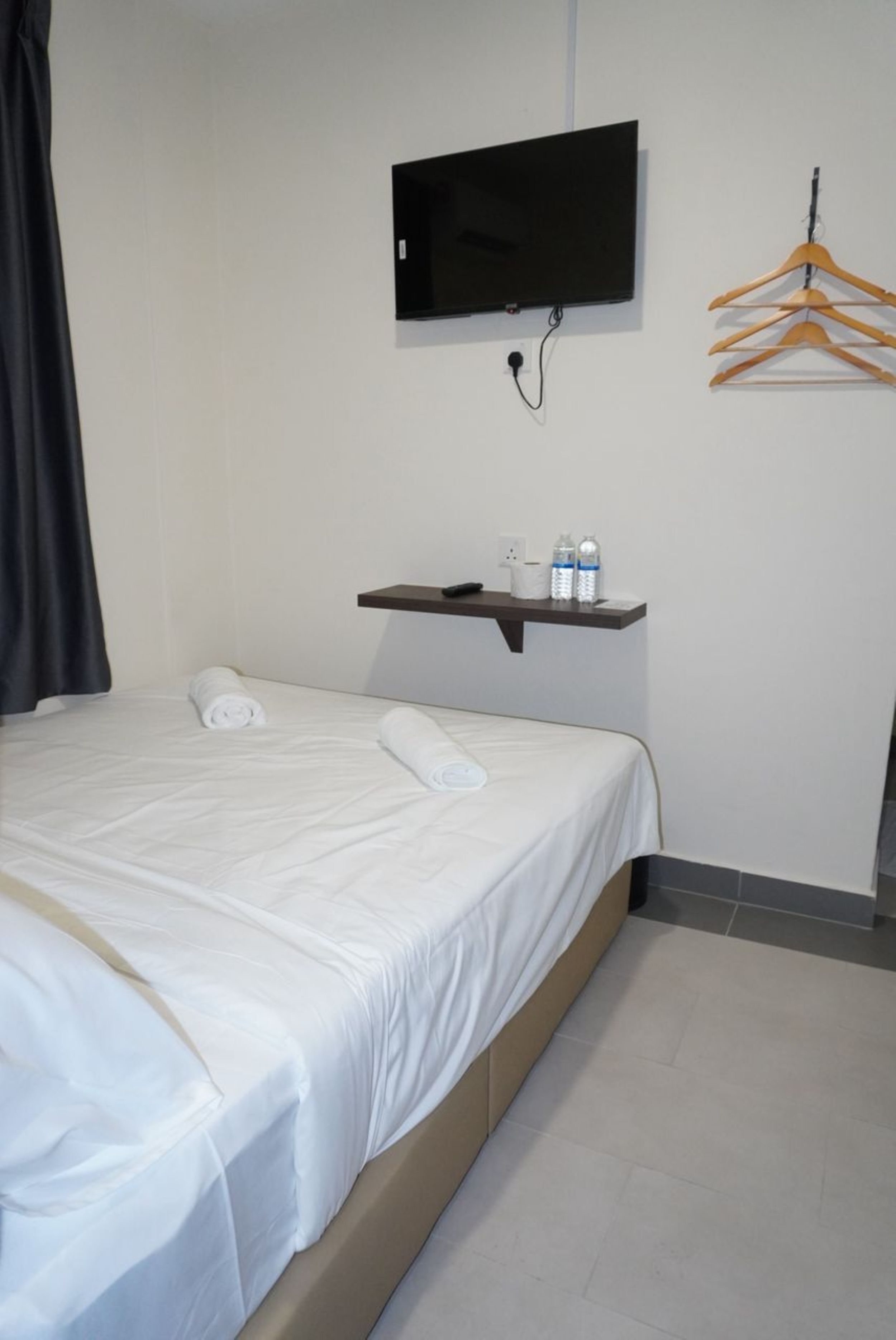 A neatly made bed with a white bedspread, a mounted television above it, a wooden hanger rack to the right, and a small shelf with bottles and a remote control.
