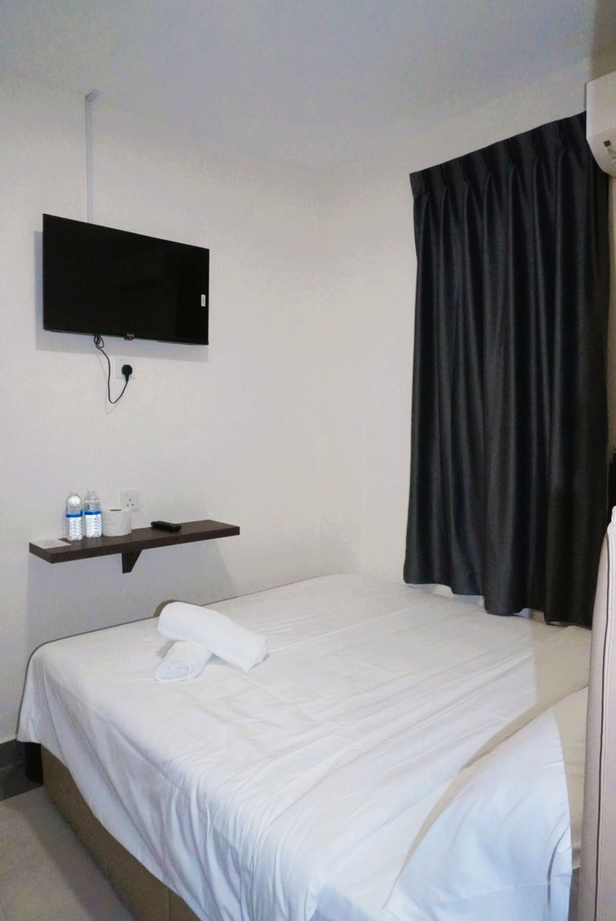Una habitación de hotel con una cama, un televisor montado, un estante con artículos de tocador y un ventanal con cortinas.