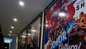 A hallway with a large poster advertising a hotel event for 2026, multiple tables, and benches along the wall. - Hotel Shlyo - Taman Nusantara Prima (Gelang Patah)