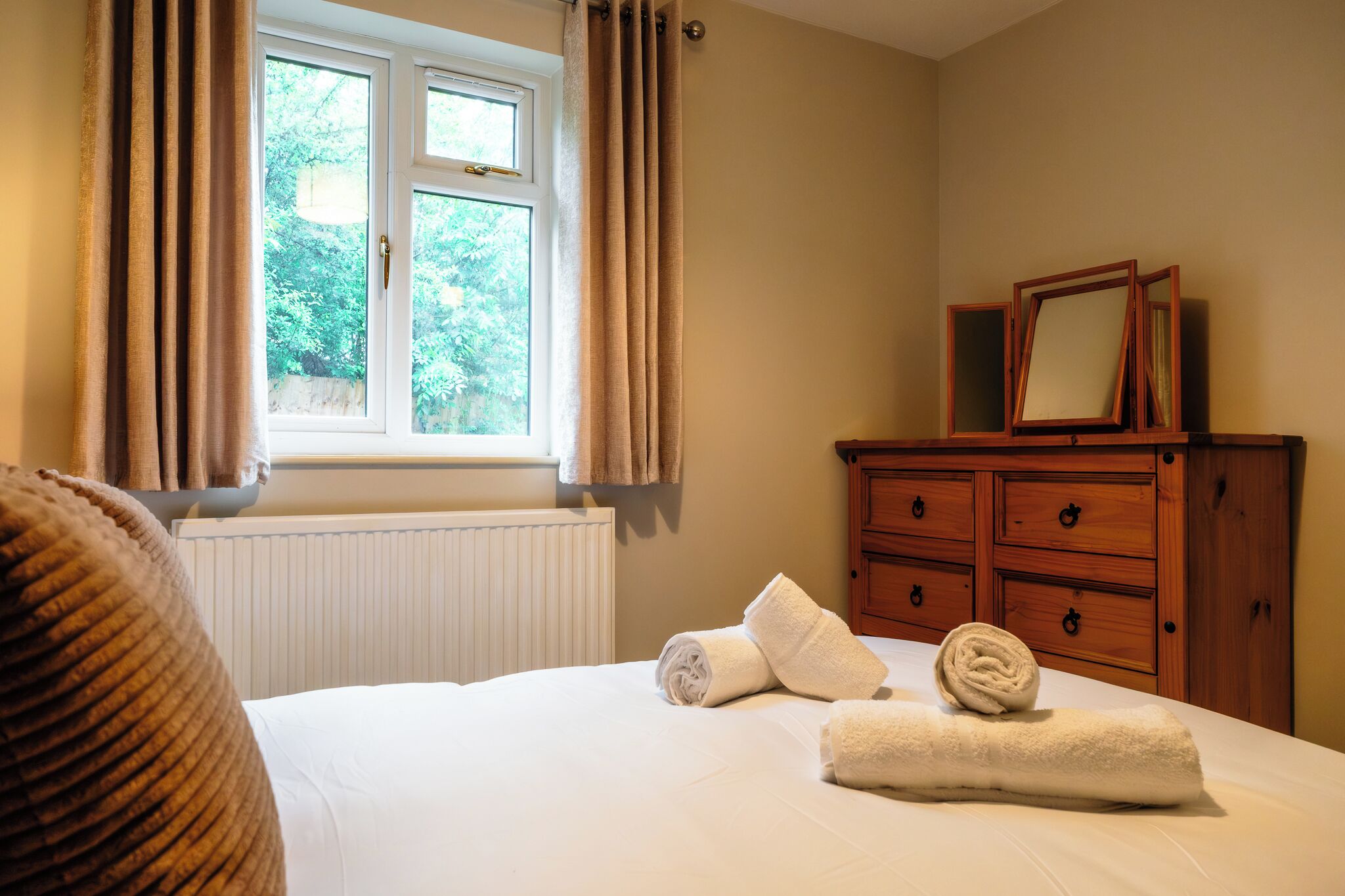 A bedroom with a bed, a window with curtains, a wooden dresser, and a mirror.