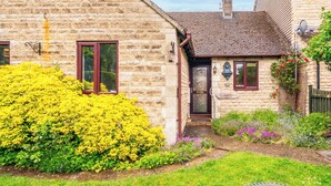 Un cottage in pietra con una porta rossa, un piccolo portico e un giardino pieno di fiori gialli e viola.