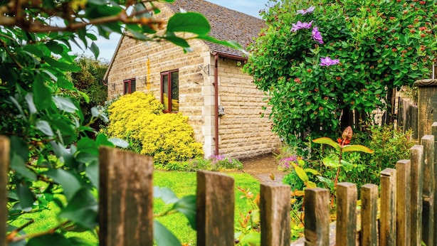 Una casa in pietra con tetto in tegole, circondata da un recinto in legno e vegetazione lussureggiante.