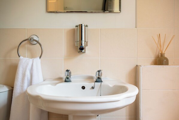 A bathroom with a white sink, two faucets, a towel ring, a mirror, and a diffuser with reed sticks.