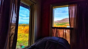 Ein Zimmer mit einem Bett, zwei Fenstern mit Vorhängen und Blick auf eine Landschaft.