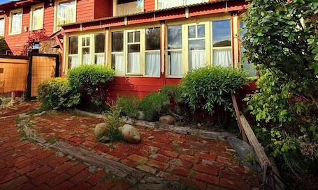 A red building with multiple windows, a brick pathway, and various green plants.. cabaña grande hasta 6 huespedes,aceptamos mascotas en San Martín de los Andes