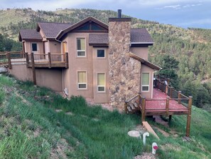 A two-story house with a stone chimney, a wooden deck, and a view of a forested hillside. - Modern Mountain Haven (Boulder)