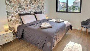 Een slaapkamer met een bed, nachtkastje, stoel en raam met uitzicht op huizen.