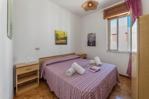 Ein Schlafzimmer mit Bett, Nachttisch, Holzkommode, Fenster mit Vorhängen und Wandbildern.