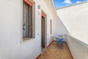Ein schmaler Korridor mit Fliesenboden, einem blauen Klapptisch mit Stuhl und einem Fenster mit dekorativer Gardine.