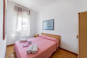 Ein Schlafzimmer mit einem Bett, einem Nachttisch, einem Fenster mit transparenten Vorhängen und einem gerahmten Bild an der Wand.