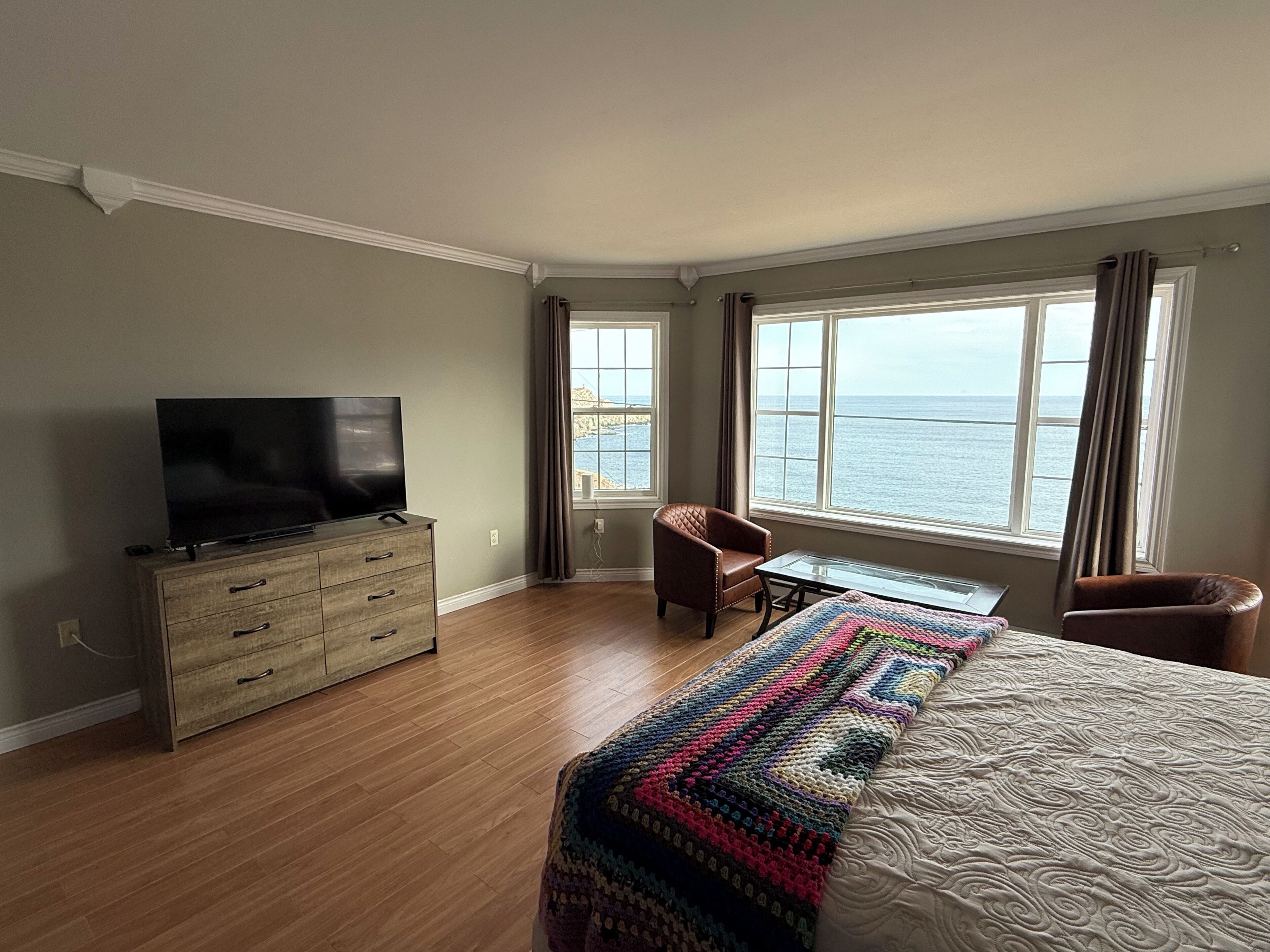 A hotel room with a television on a dresser, a chair by the window, a bed with a colorful blanket, and a view of the sea.. Oceanfront retreat with stunning coastal views and tranquility