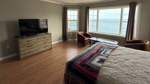 Una camera d'albergo con televisione sul comodino, una sedia vicino alla finestra, un letto con coperta colorata e vista sul mare.