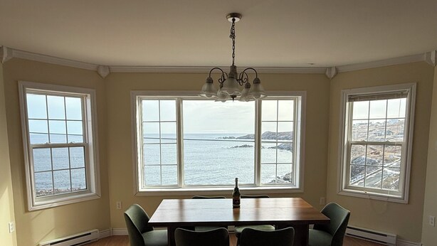 Zona pranzo con tavolo e sedie in legno, ampie finestre vista mare e un lampadario.
