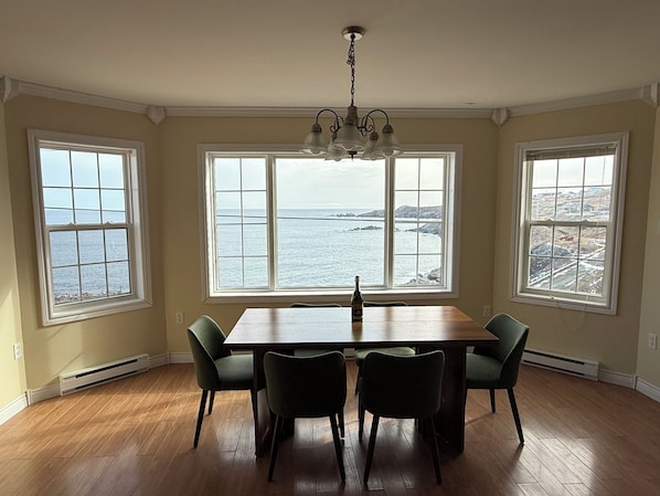 A dining area with a wooden table and chairs, large windows with a view of the sea, and a chandelier.