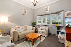 A living room with a sofa, armchair, coffee table, and a window with shutters. - Tranquil Cottage | Lovely Street, Walk to Leura (Leura)