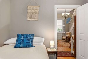 A bedroom with a bed, a hanging macrame wall decor, a blue pillow, a nightstand with a lamp, and a view into a hallway with a chair and a staircase. - Tranquil Cottage | Lovely Street, Walk to Leura (Leura)