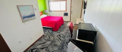 A compact hotel room with a bed, a desk with a computer, a window, and a door.