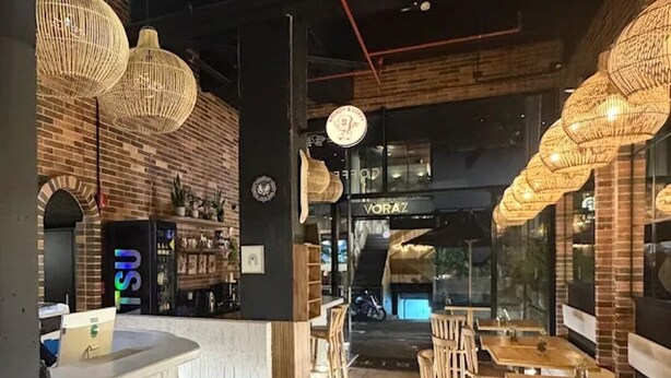 Een restaurantinterieur met een receptiebalie, houten stoelen, hangende rieten lampen en een bakstenen muur.