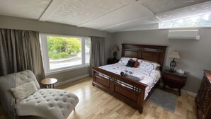 Ein Schlafzimmer mit einem großen Bett, einem Sessel, einem Nachttisch, einer Lampe und einem Fenster mit Blick auf Grünfläche.