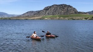 Zwei Personen paddeln auf einem ruhigen See vor einer gebirgigen Kulisse.