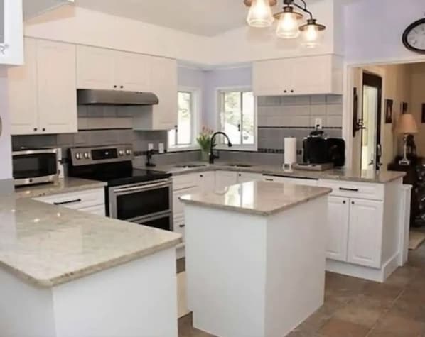 Une cuisine avec des meubles blancs, un îlot central, des appareils en acier inoxydable et des luminaires suspendus.
