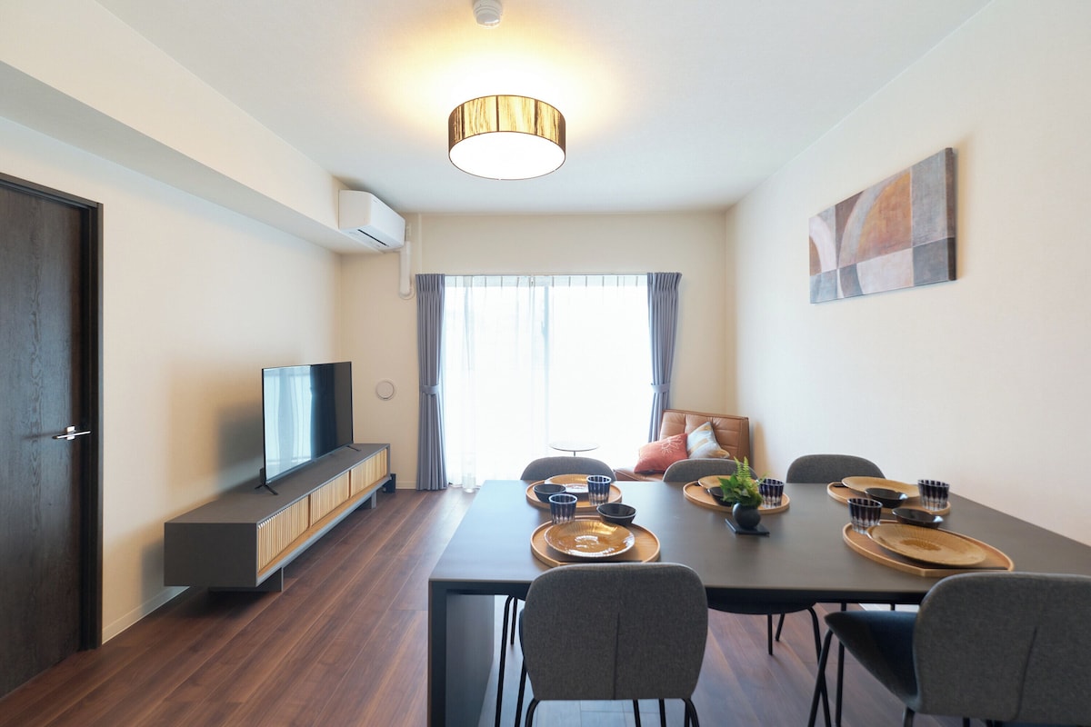 A modern living room with a flat-screen TV, a dining table set for four, and a sofa in the background.