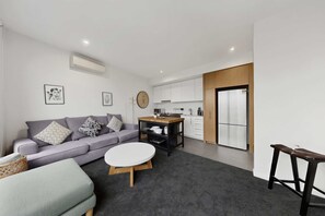 Ein modernes Wohnzimmer mit einem grauen Sofa, einem runden Couchtisch und einer Küchenzeile mit weißen Schränken und einem Kühlschrank.