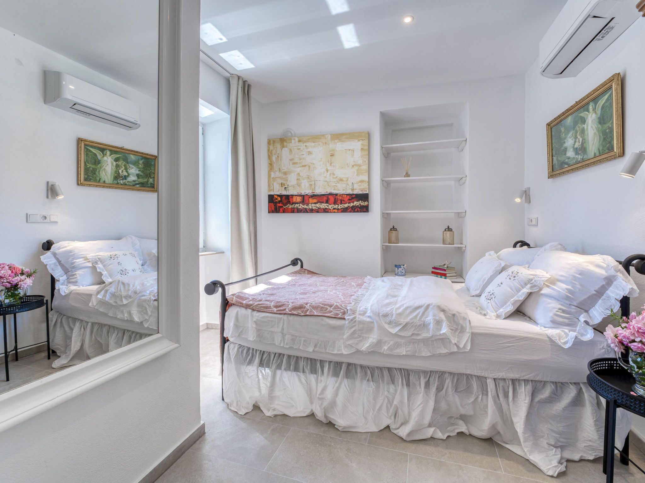 A bedroom with a bed, a bedside table with flowers, a painting, and a shelf with decorative items.